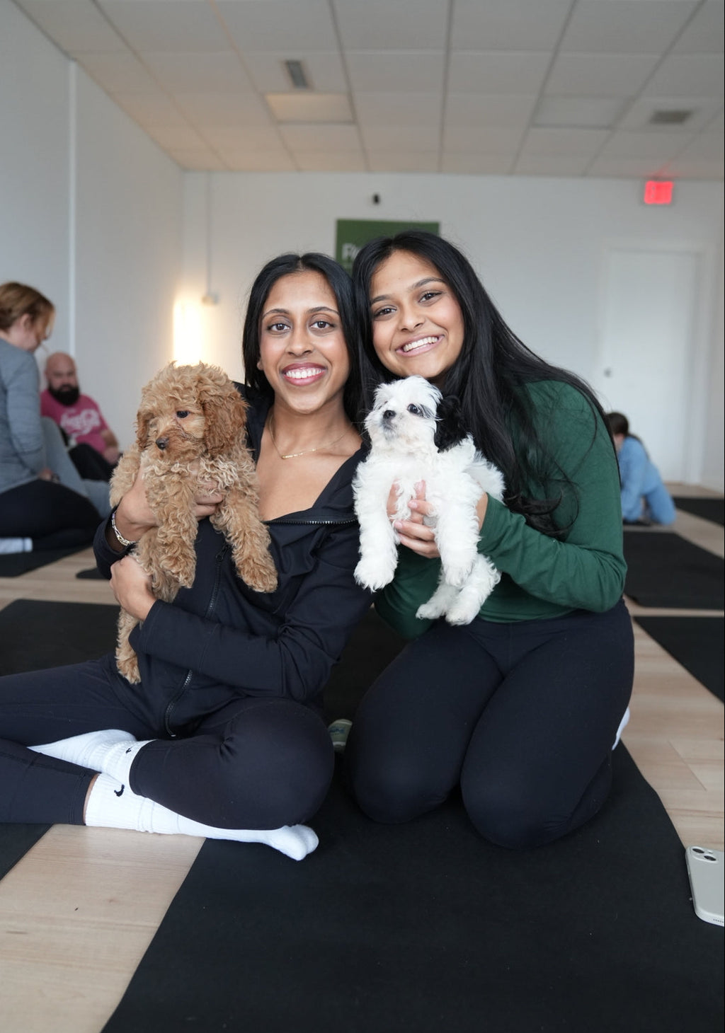 Two women sitting on a yoga mat with two small dogs, smiling at the camera during puppy yoga by Reset Vibes